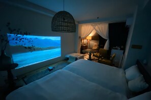 A hotel room with a large bed, a small seating area, a TV screen displaying a scenic view, and a hanging light fixture.