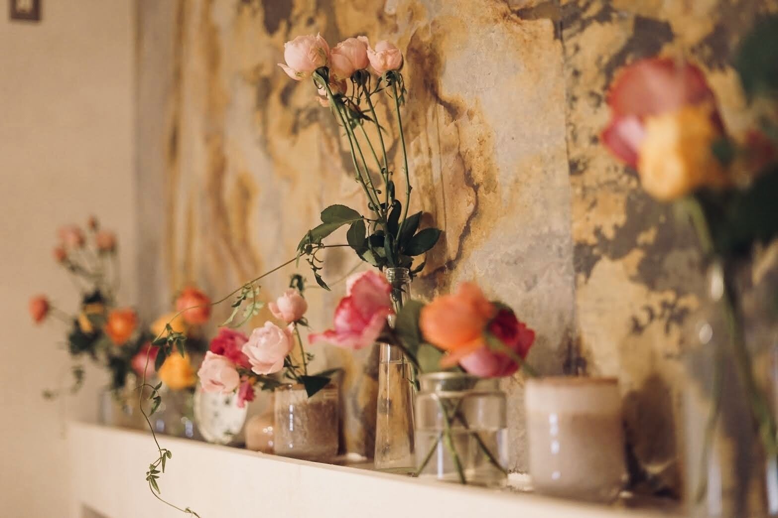 A shelf with various glass vases containing flowers, set against a textured wall.