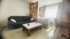 A hotel room with a dark-colored sofa, a white coffee table, a bed with white bedding, a wooden partition with decorative patterns, and a desk with a chair.