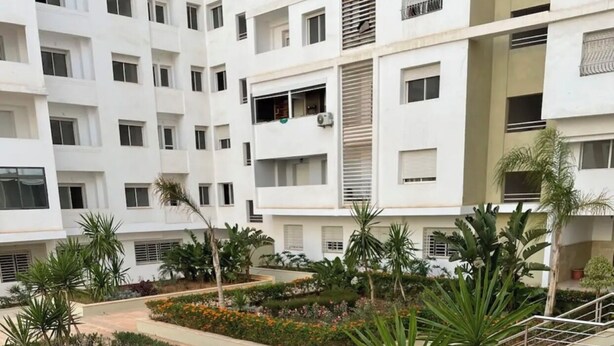Un edificio residencial de varios pisos con balcones, un jardín ajardinado con diversas plantas y flores, y una barandilla de metal.