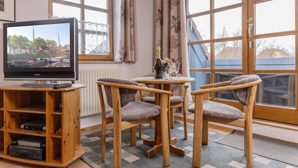 A wooden TV stand with a flat-screen TV, two upholstered chairs, a small round table with a bottle and flowers, and large windows with curtains. - Einfach mal Ausspannen: Die Obergeschoss-wohnung Liegt in Unmittelbarer Nähe ein (Carolinensiel)
