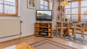 A living room with a television on a wooden stand, a dining area with two wooden chairs, and large windows with curtains. - Einfach mal Ausspannen: Die Obergeschoss-wohnung Liegt in Unmittelbarer Nähe ein (Carolinensiel)