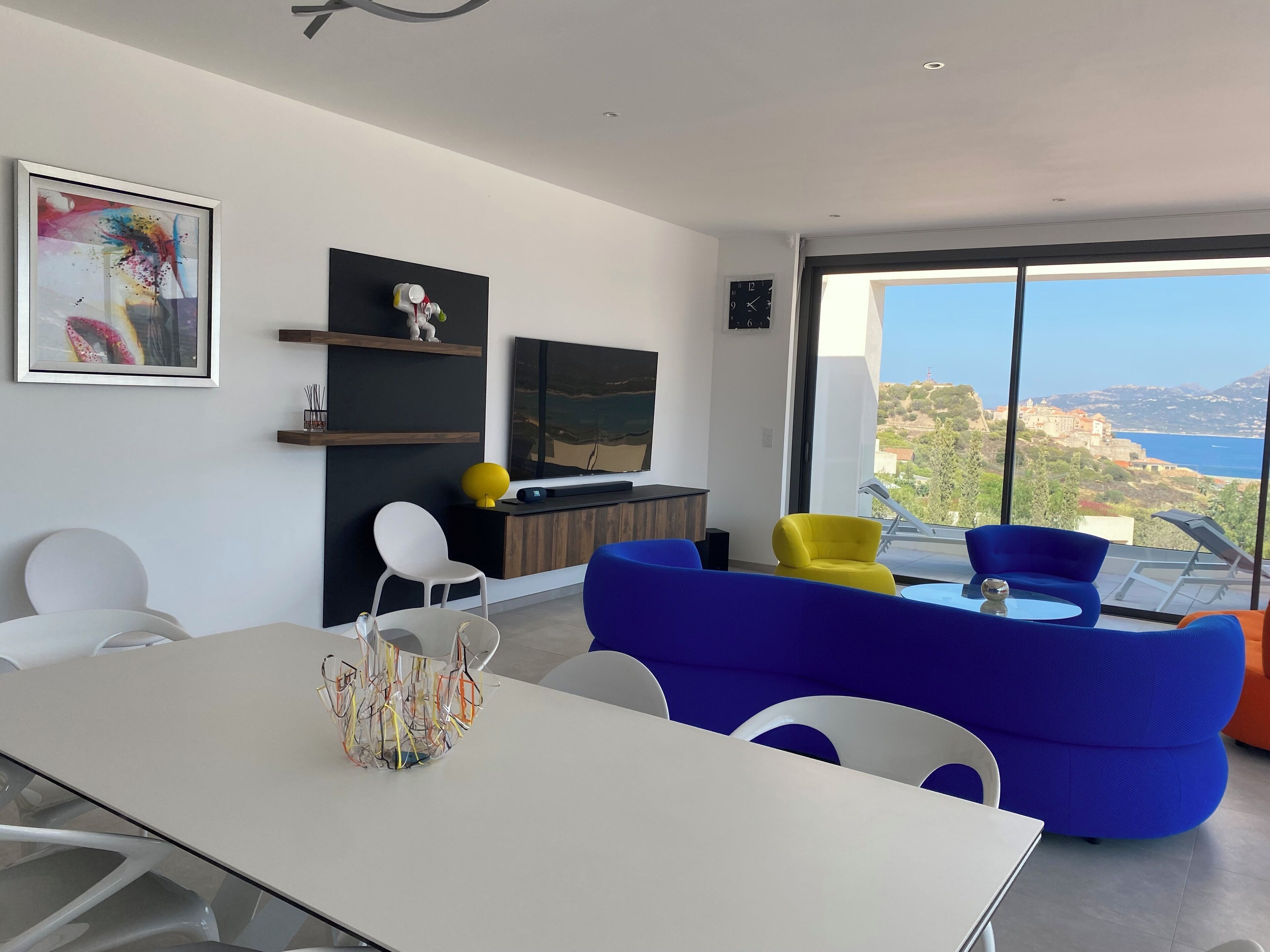 Una sala de estar moderna con comedor, un ventanal con vista al mar y un televisor montado.