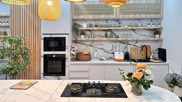 A modern kitchen with a built-in oven, a microwave, and a gas stove. There are hanging wicker lamps, marble shelves with kitchenware, and a potted plant. - Herma Hotel (Bang Phli)