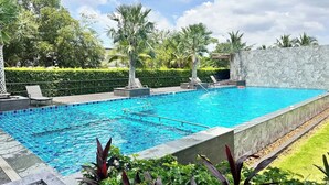 A rectangular swimming pool with clear blue water, surrounded by a stone wall and lush greenery, including palm trees and well-maintained hedges. - Herma Hotel (Bang Phli)