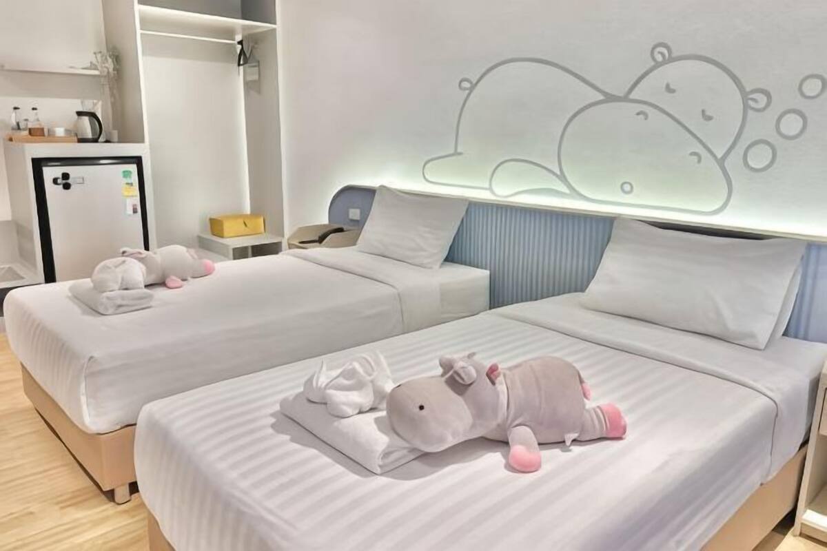 Two beds with white bedding, a bedside table, a nightstand, a wall-mounted light, and a refrigerator.