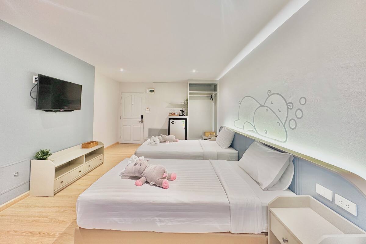 A modern hotel room with two beds, a flat-screen TV, a small kitchenette, and a bathroom visible through an open door.