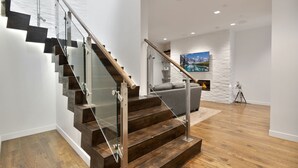 A modern staircase with glass railings and wooden steps, leading to a living area with a fireplace and a painting of a mountain landscape.