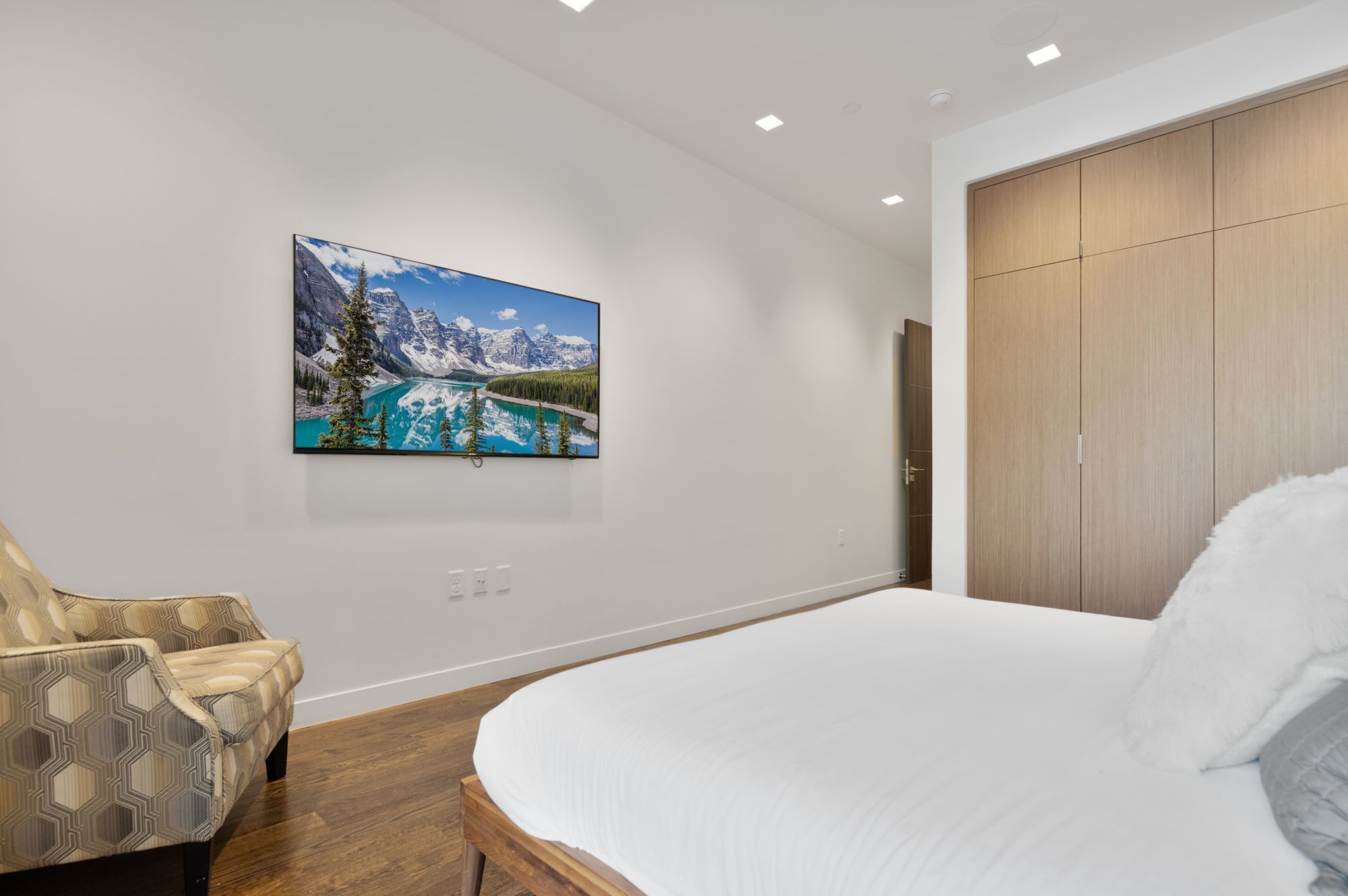 A bedroom with a large bed, a chair, a wardrobe, and a wall-mounted painting of a mountain landscape.