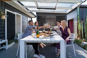 A group of people toasting at a white table with a variety of food and drinks, set on a wooden deck with plants and a modern building in the background. - Maggie’s the Little Black Cottage - In the Heart of the Hunter Valley (Cessnock)