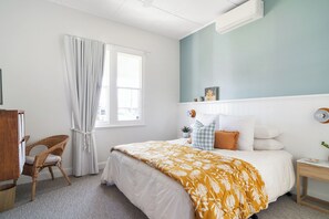 A bedroom with a bed, a chair, a window with curtains, a bedside table, and a wall-mounted air conditioner. - Maggie’s the Little Black Cottage - In the Heart of the Hunter Valley (Cessnock)