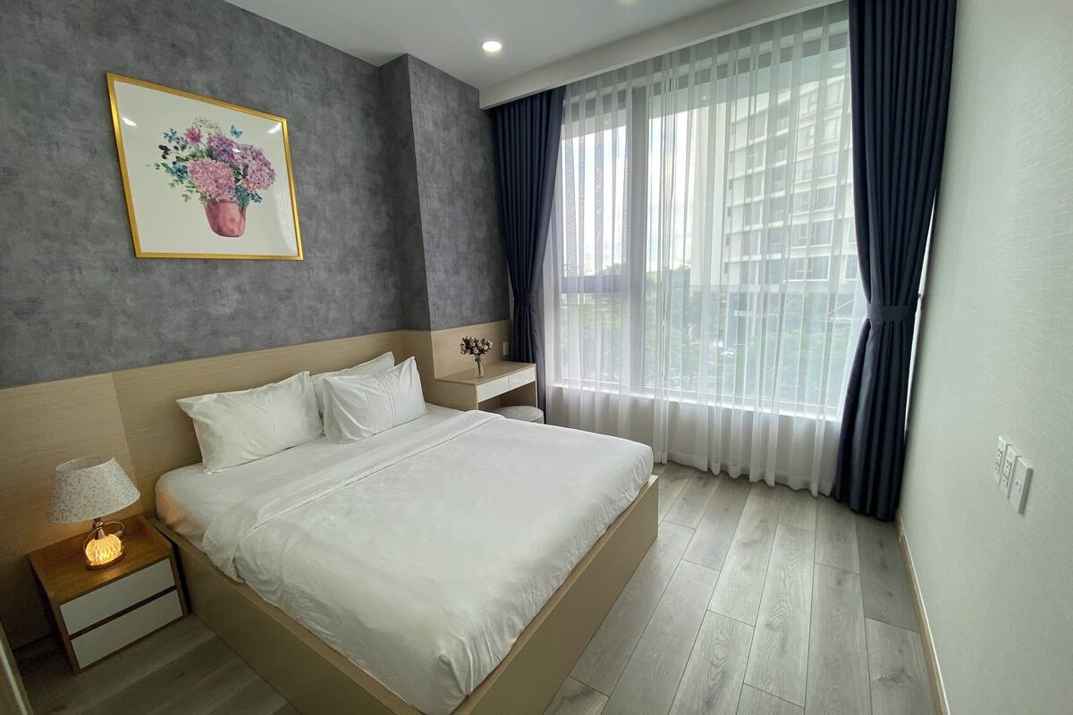 Habitación de hotel moderna con una cama grande, mesitas de noche, una ventana con cortinas traslúcidas y una obra de arte floral enmarcada en la pared.