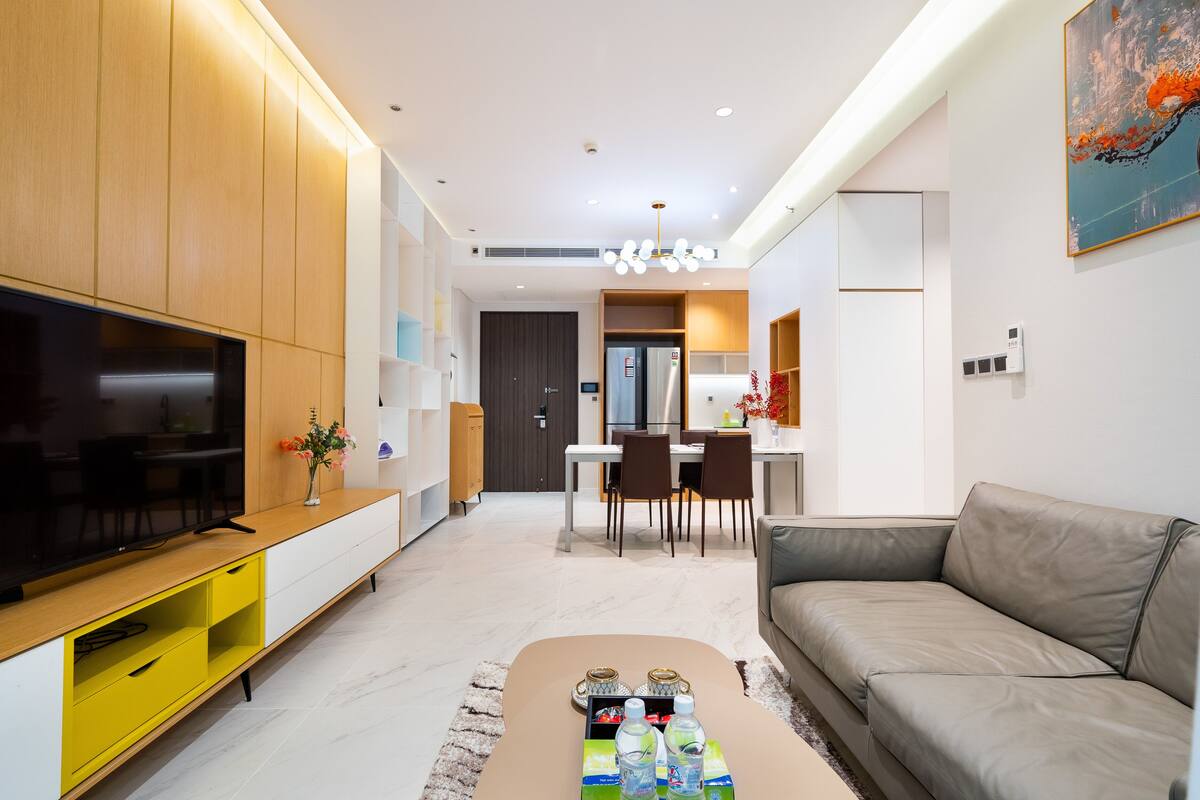 Una sala de estar moderna con televisor de pantalla plana, un mueble de almacenaje amarillo, sofá color beige y un comedor al fondo.