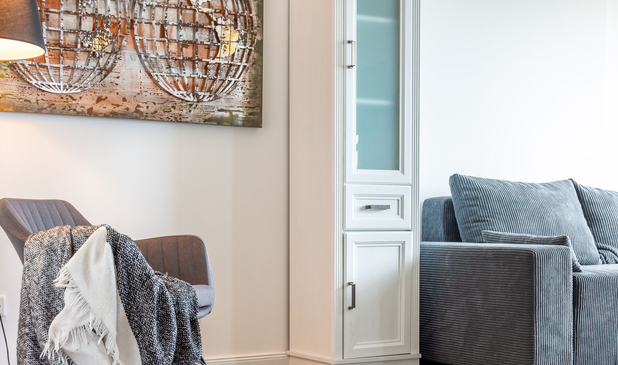 Una habitación con un cuadro abstracto grande, un sillón gris, un sofá azul y un mueble blanco.