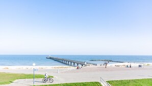 A coastal promenade with a long pier extending into the sea, a bicycle path, and a grassy area with a bench.