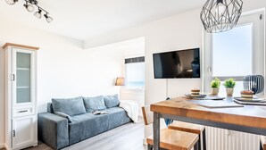 A modern living room with a sofa, a dining table with chairs, a TV mounted on the wall, and a window with a view.