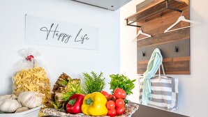 A kitchen with a 'Happy Life' sign, a basket of fresh vegetables, a bag of pasta, and a striped bag on the counter.