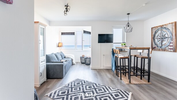A modern living room with a sofa, a dining area with bar stools, a kitchen with a TV, and a large window with a view.