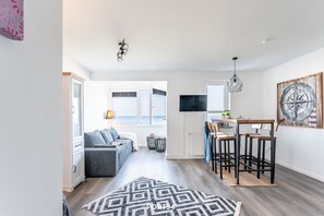 Uma sala de estar moderna com sofá, área de jantar com banquetas, cozinha com televisão e uma janela grande com vista.