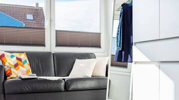 A black leather sofa with colorful pillows, a white throw, and a window with blinds.