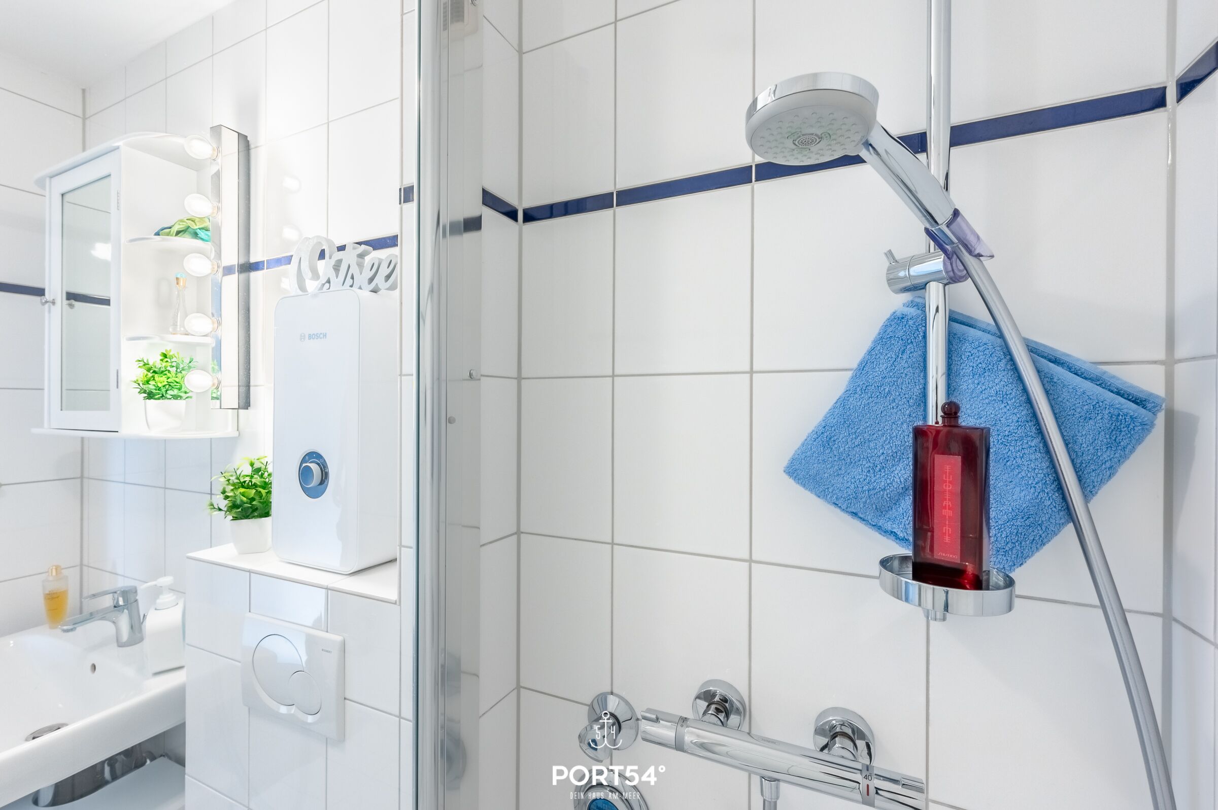 A modern bathroom with a white sink, mirror with lights, Bosch water heater, shower with blue towel, and PORT54° water heater.