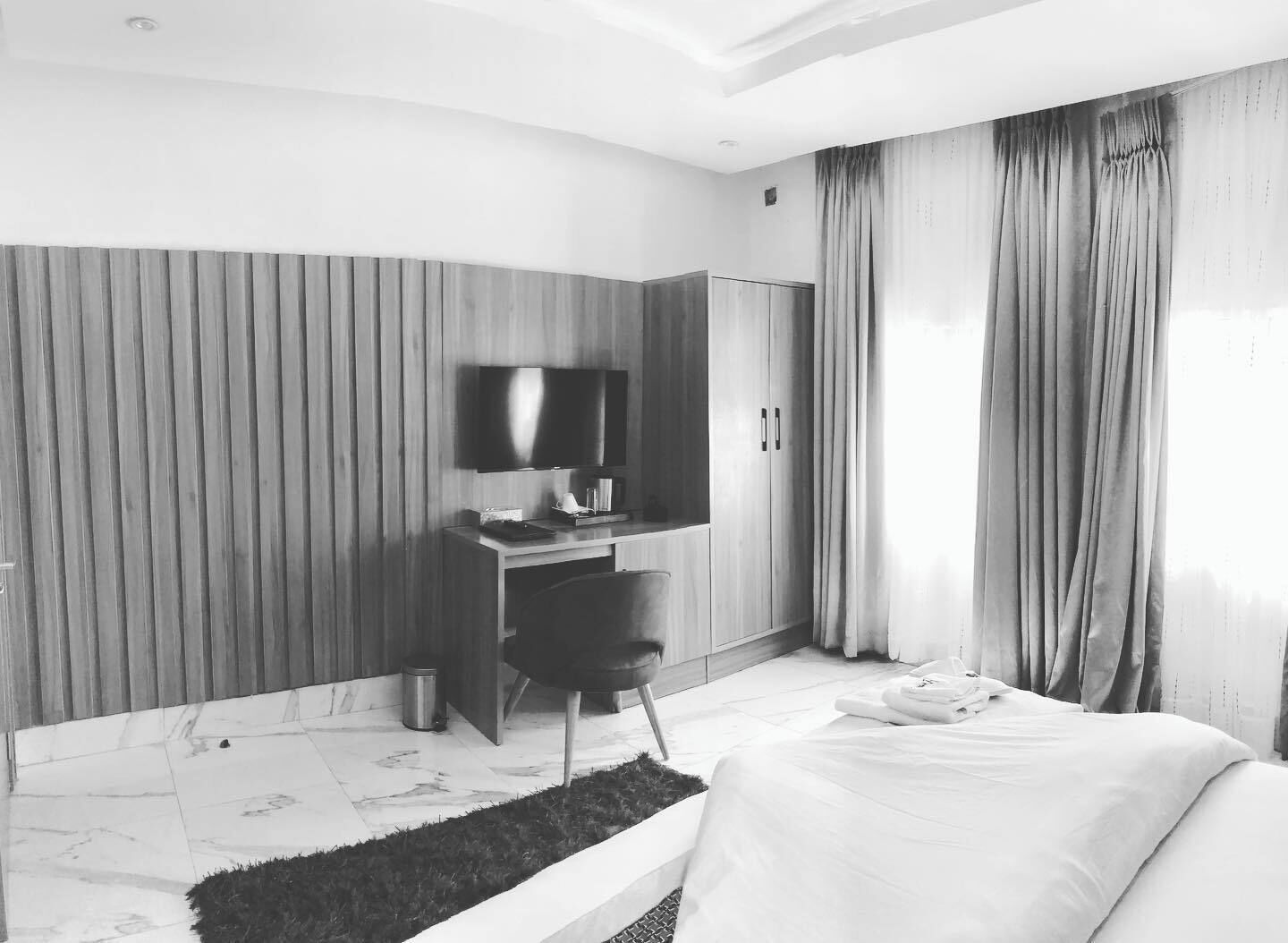 A hotel room with a bed, a desk with a flat-screen TV, a wardrobe, and a large window with curtains.