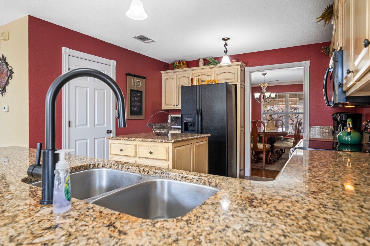 Una cocina con encimeras de granito, pileta de acero inoxidable, refrigerador negro y alacenas de madera.