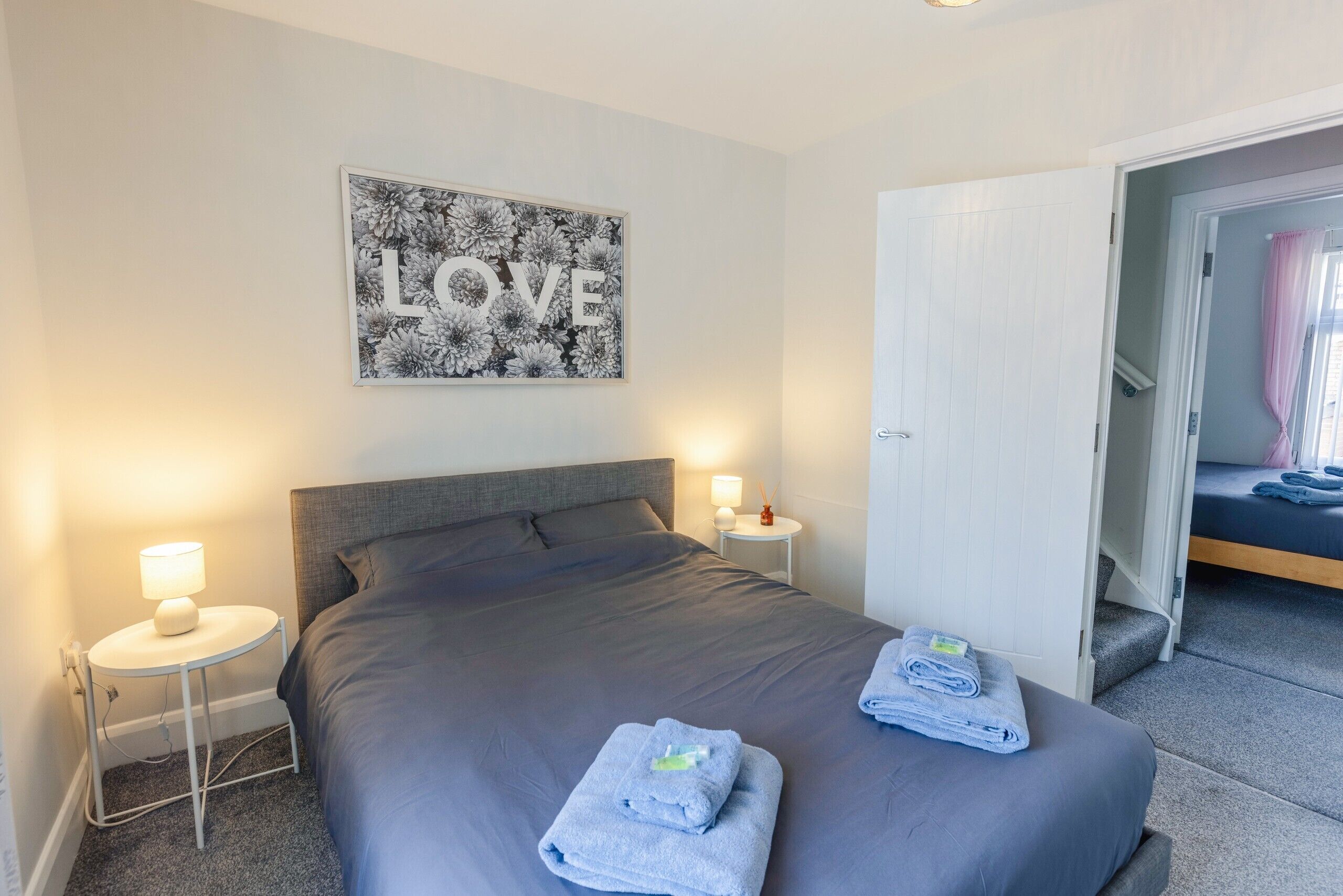A neatly made bed with a grey headboard, two bedside tables with lamps, and a wall art piece with the word "LOVE".