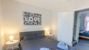 Un lit bien fait, avec une tête de lit grise, deux tables de chevet avec des lampes et une œuvre d’art murale sur laquelle est inscrit le mot « AMOUR ».