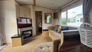 A cozy RV interior with a TV, sofa, and a window with curtains. - Burgh Castle 3-Bed Haven: Near Norfolk Broads (Burgh Castle)