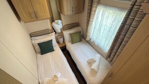 A compact bedroom with two single beds, a small bedside table, a cabinet above the beds, and a window with curtains. - Burgh Castle 3-Bed Haven: Near Norfolk Broads (Burgh Castle)