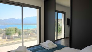 Ein Hotelzimmer mit einer großen Glasschiebetür, die auf einen Balkon mit Blick auf einen See und Berge führt. Es gibt ein Bett mit weißer Bettwäsche und einer blauen Tagesdecke.