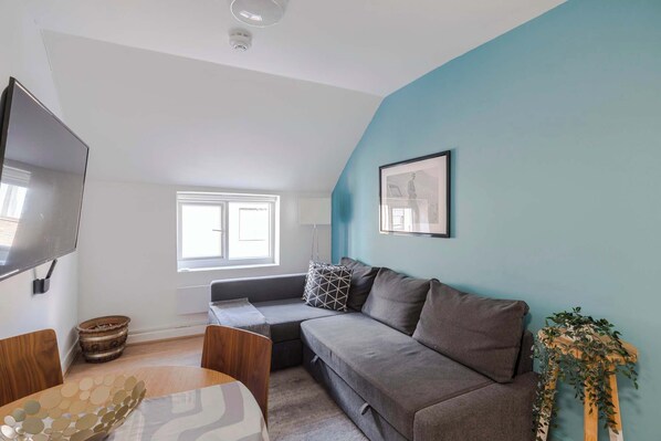 A modern living room with a grey sofa, a wooden coffee table, a TV mounted on the wall, and a window providing natural light.