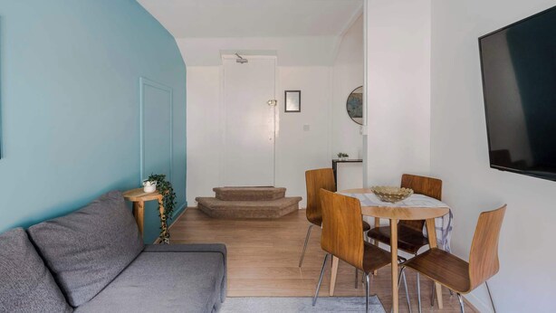 A modern living room with a grey sofa, a dining area with wooden chairs and a small table, and a TV mounted on the wall. - Quiet Mews 1-Bed · Fitzrovia · Sleeps 4 (Greater London)