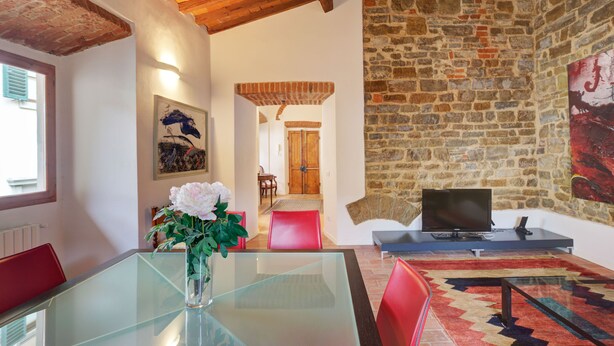 A modern living room with a glass table, red chairs, a stone wall, and a television.