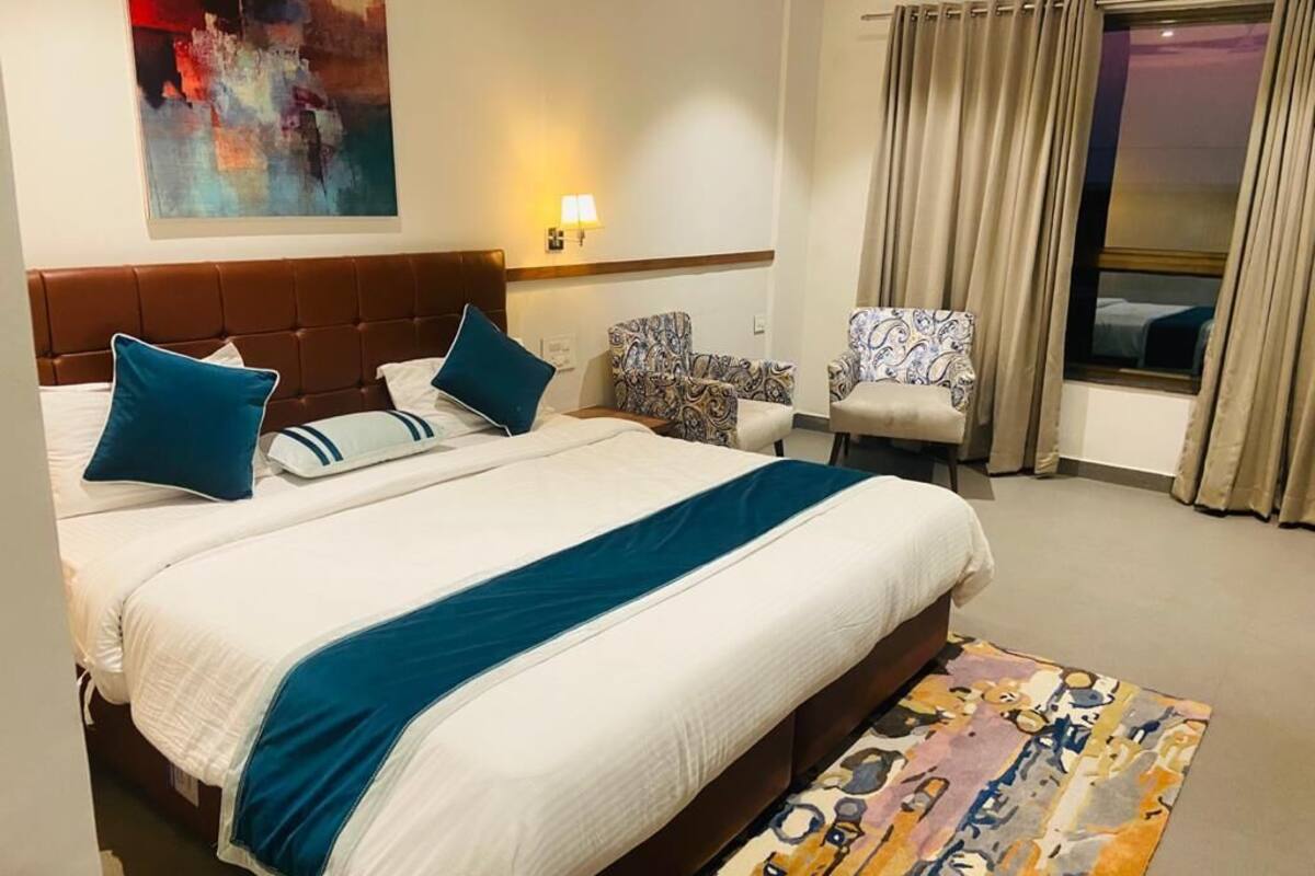 A hotel room with a large bed, two chairs, a window with curtains, and a painting on the wall.