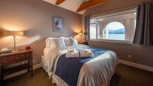 A bedroom with a large bed, a nightstand, a desk, a window with a view, and a painting on the wall. - Glacier + Tide Retreat (Homer)