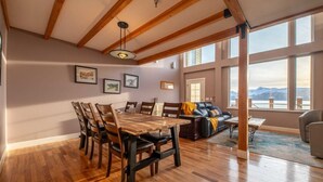 A dining area with a wooden table and chairs, a sofa, and a view of the mountains through large windows. - Glacier + Tide Retreat (Homer)
