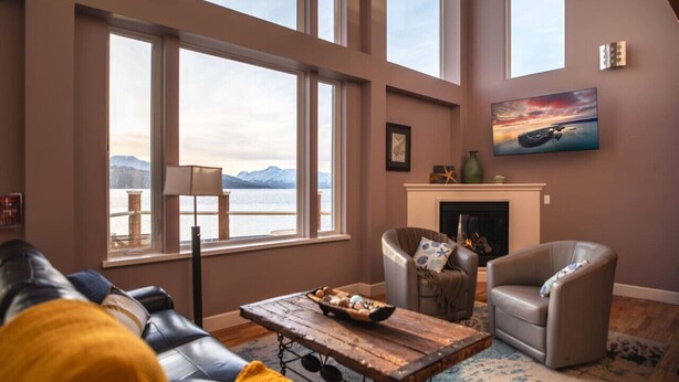 A living room with a fireplace, a large window with a view of mountains, and a cozy seating area. - Glacier + Tide Residence (Homer)