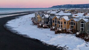 A coastal area with snow-covered buildings, a beach, and a distant shoreline. - Glacier + Tide Residence (Homer)