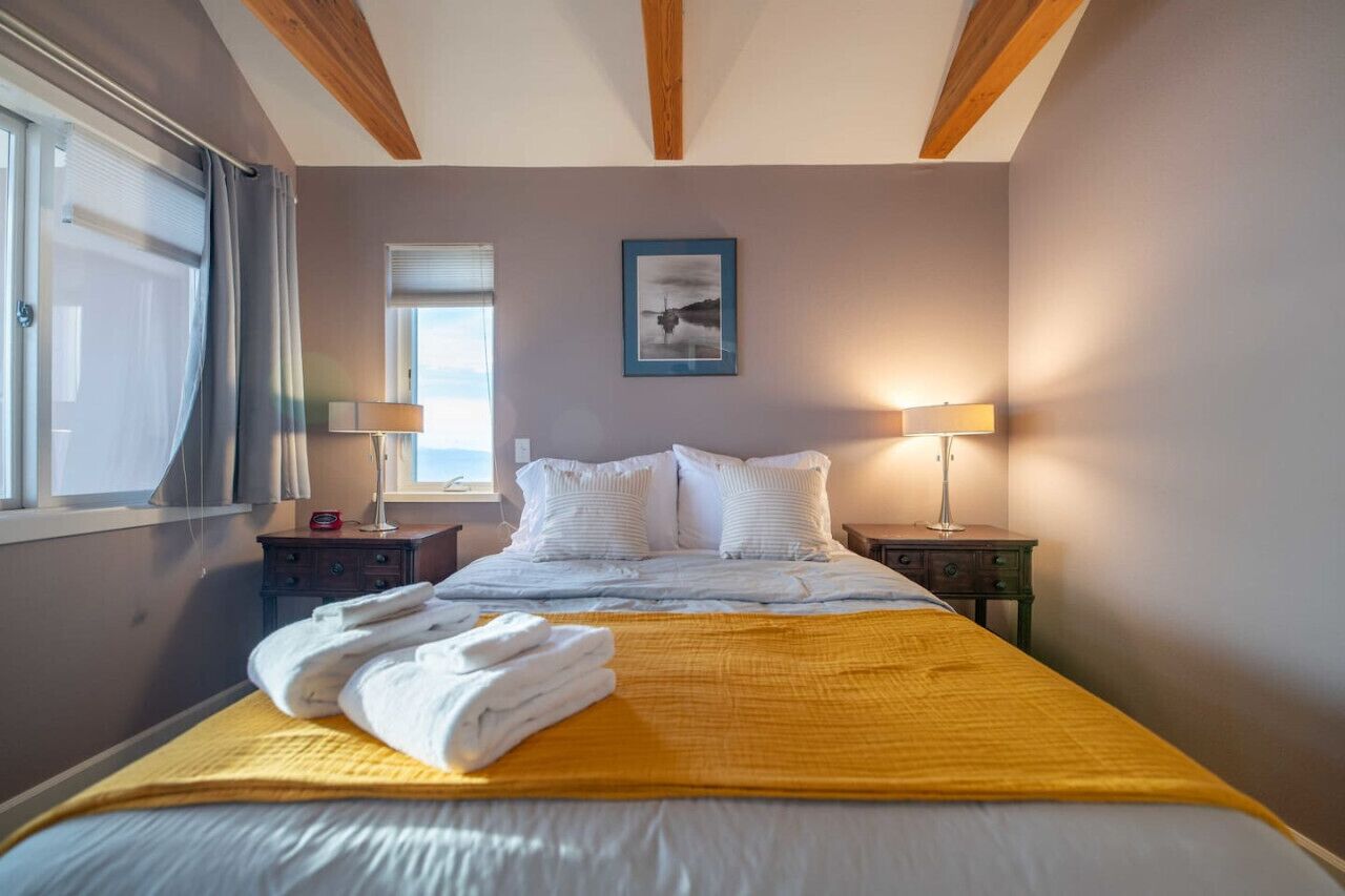 Ein Hotelzimmer mit einem Bett, zwei Nachttischen, einem fenster mit Vorhängen, einem gerahmten Bild an der Wand und einer Lampe auf jedem Tisch.