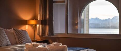 A hotel room with a bed, a nightstand with a lamp, a window with a view of mountains, and towels on the bed.