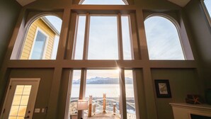 A room with a large window overlooking a body of water and mountains, a door, and a fireplace. - Glacier + Tide Residence (Homer)