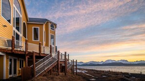 A yellow building with a balcony, a staircase, and a view of mountains and water. - Glacier + Tide Residence (Homer)