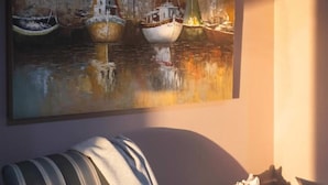 A living room with a striped armchair, a small round table with a decorative bowl, and a large painting of boats on the wall. - Little Glacier + Tide (Homer)