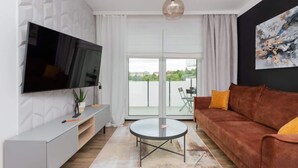 Een moderne woonkamer met een flatscreen televisie, een bruine bank, een glazen salontafel en een groot raam met uitzicht op groen.