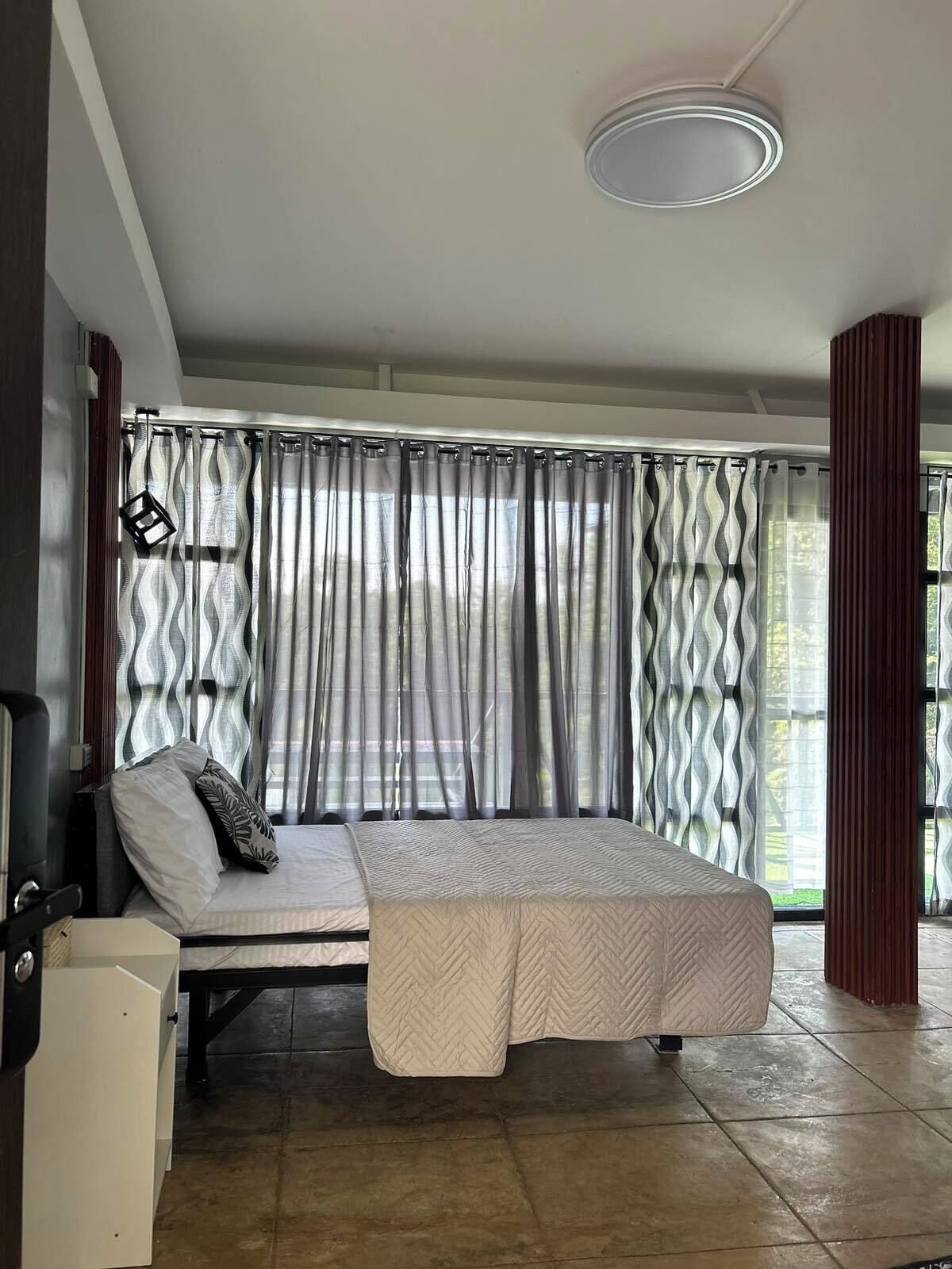 Una stanza con un letto, un tavolino e una sedia. Si vede una finestra con tende, una luce a soffitto e una porta.
