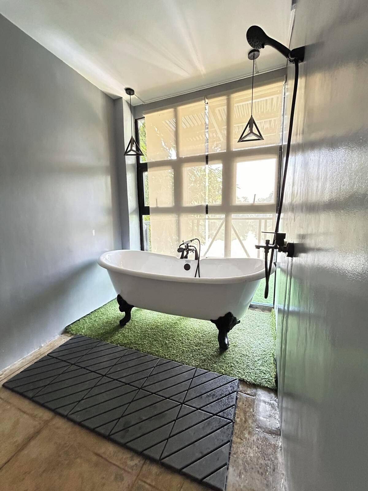 A bathroom with a freestanding bathtub, a window with blinds, and a green artificial grass floor.