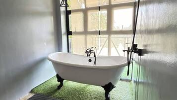 A bathroom with a freestanding bathtub, a window with blinds, and a green artificial grass floor.