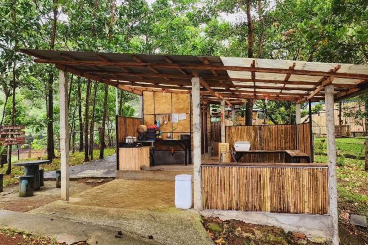 Una estructura de madera rústica con un área de estar cubierta, una pequeña cocina y un sendero de concreto.